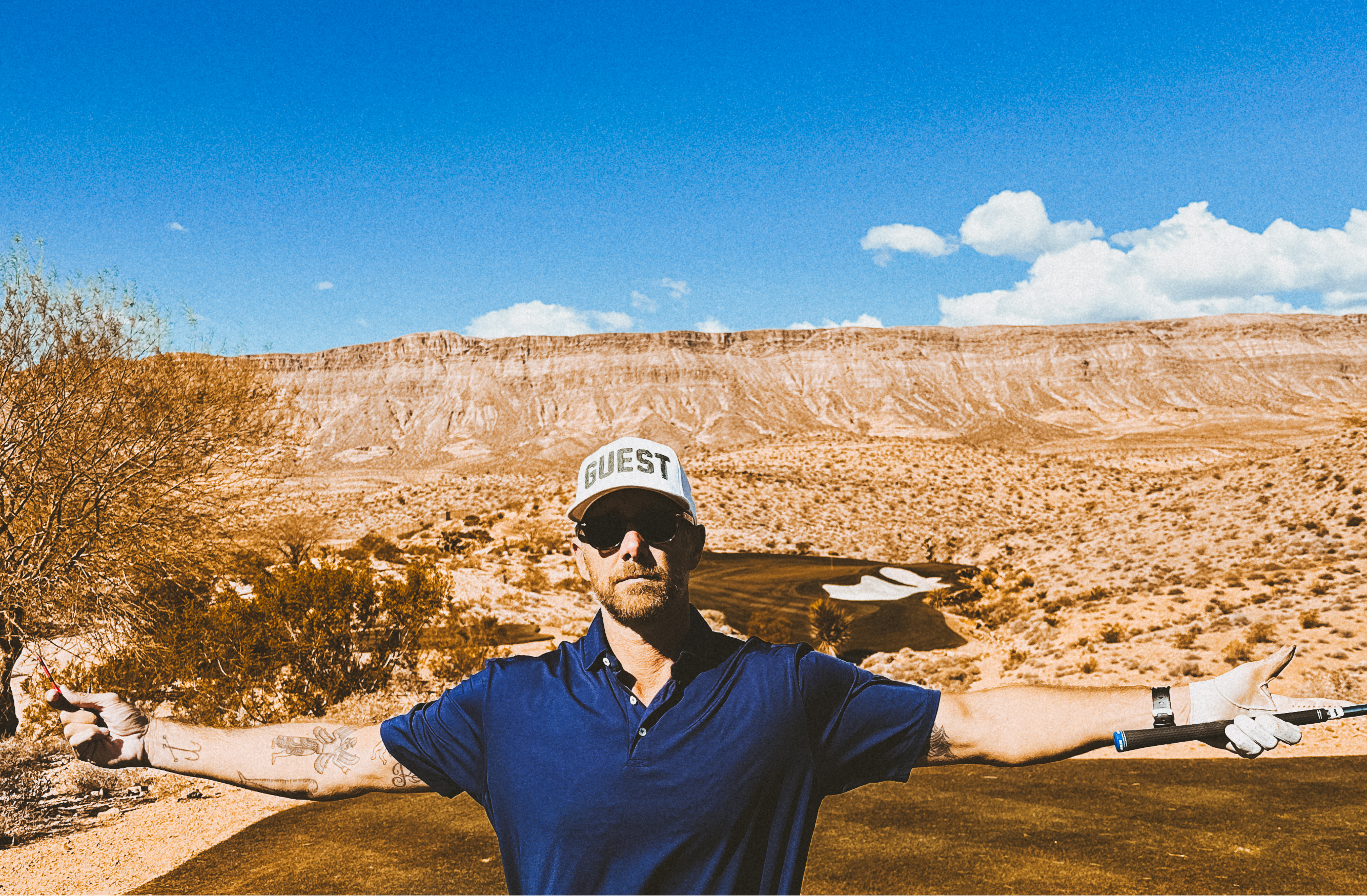 Golfer standing on a desert golf course with arms outstretched, wearing sunglasses and a white country club guest hat