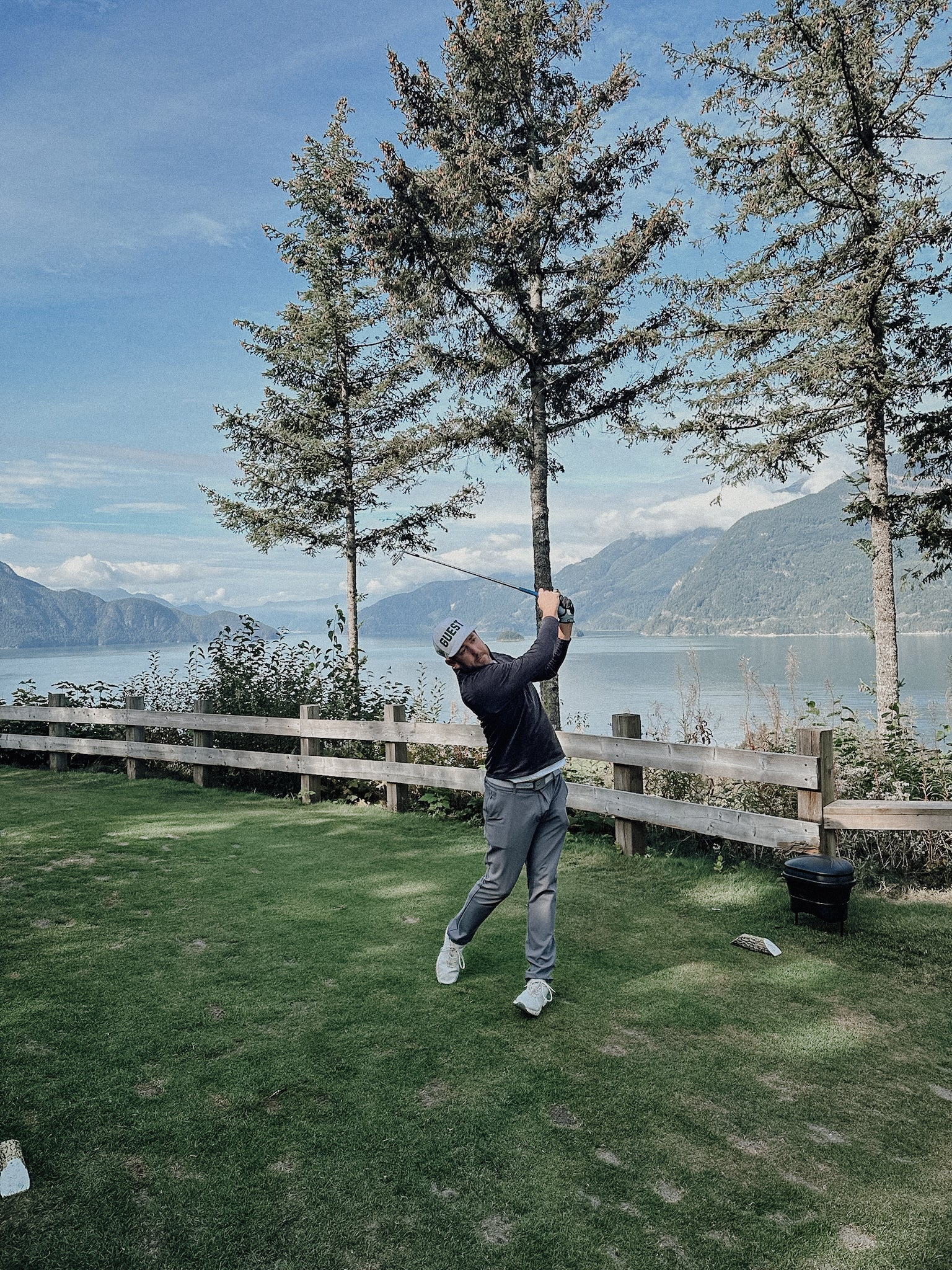 Golfer wearing a classic country club guest hat taking a swing on a lakeside golf course with mountains in the background