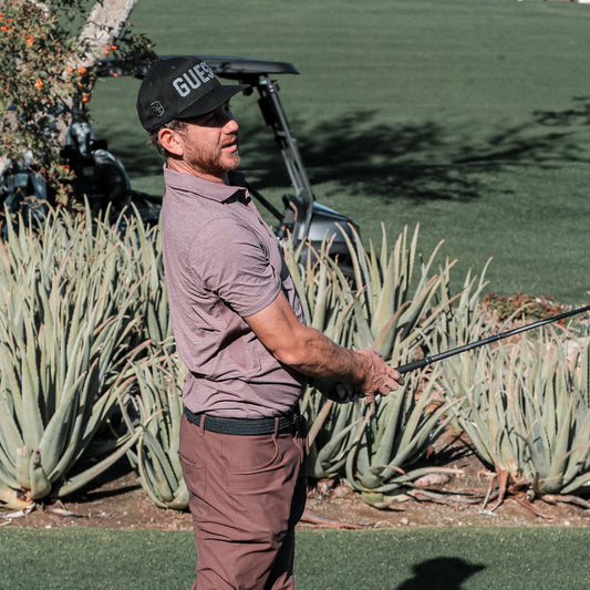 Golfer wearing a twilight Country Club GUEST hat swinging a club on a sunny golf course surrounded by desert plants