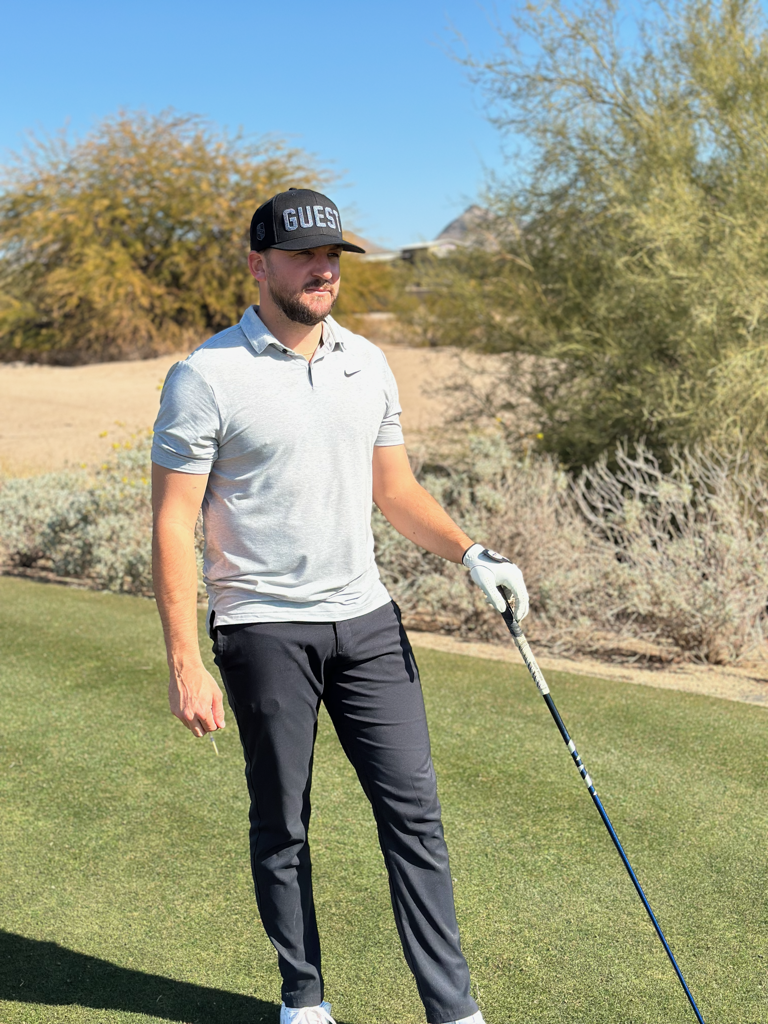 Golfer standing on the tee box holding a club and wearing a twilight country club guest hat with desert scenery in the background