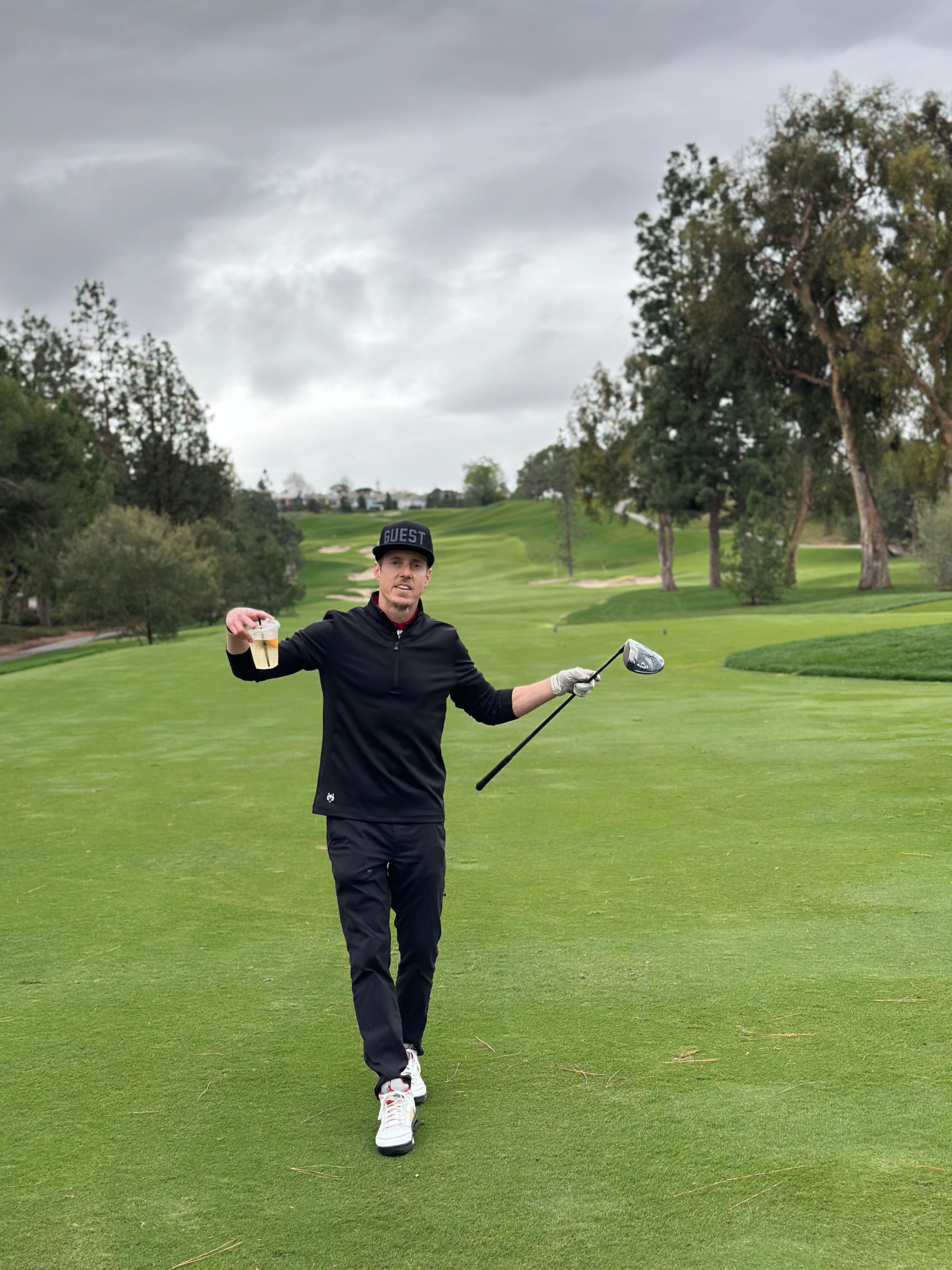 Golfer walking down the fairway holding a drink and a driver wearing a twilight country club guest hat on an overcast day