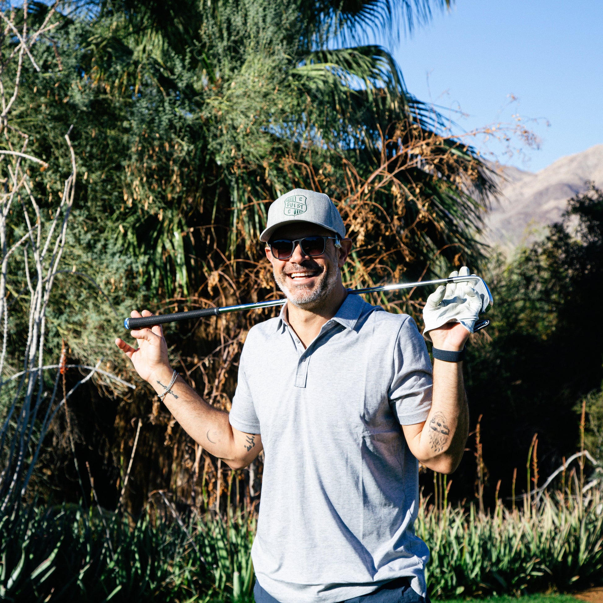 Smiling golfer holding a club over his shoulders while wearing a gray country club guest hat